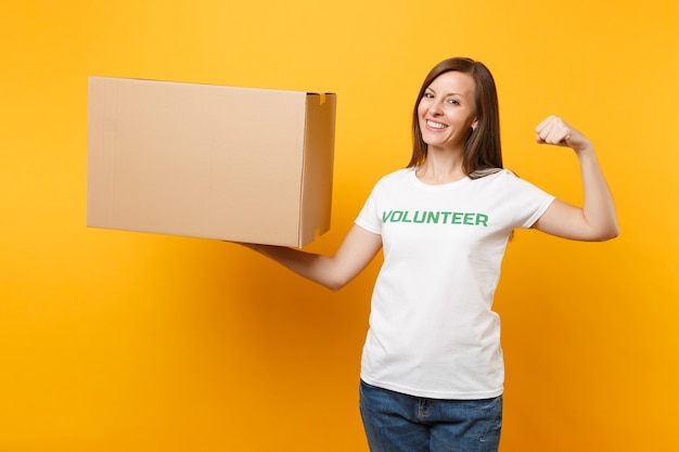 Retrato de mujer sonriente en camiseta blanca con inscripción escrita voluntaria de título verde con caja de cartón grande aislada sobre fondo amarillo. Ayuda de asistencia gratuita voluntaria, concepto de gracia de caridad.