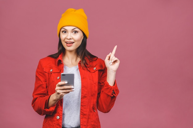 Retrato de una mujer sonriente casual alegre que sostiene el teléfono móvil y que señala el dedo lejos aislado sobre la pared rosada.