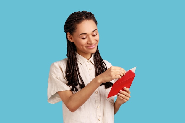 Foto retrato de una mujer sonriente con dreadlocks negros de pie sobre rojo abierto con felicitaciones, leyendo una carta romántica, usando pantalones blanco. disparo de estudio interior aislado sobre fondo azul.