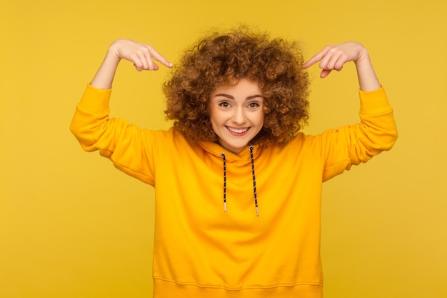 Foto retrato de una mujer sonriente emocionada con capucha de estilo urbano que señala su peinado afro moderno, cosméticos publicitarios, acondicionador o champú para el cabello rizado. tiro de estudio interior aislado sobre fondo amarillo