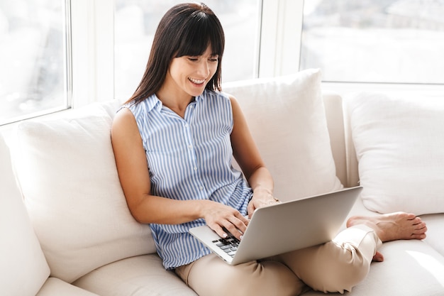 Retrato de mujer sonriente escribiendo y usando la computadora portátil plateada mientras está sentado en el sofá en el apartamento luminoso