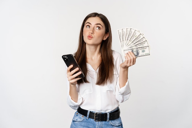 Retrato de una mujer sonriente feliz usando una aplicación de teléfono móvil, sosteniendo dinero en efectivo, de pie sobre fondo blanco