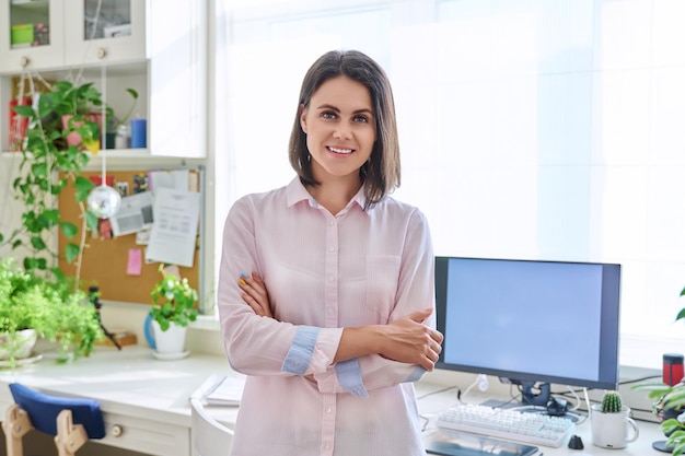 Retrato de mujer sonriente joven mirando a la cámara de pie en la oficina en casa