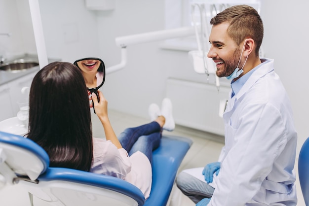 Retrato de una mujer sonriente mirándose al espejo checando los dientes después de un procedimiento dental en la clínica