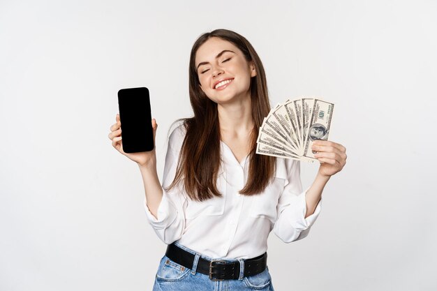 Foto retrato de mujer sonriente mostrando la pantalla del teléfono móvil y dinero en efectivo, luciendo feliz, de pie sobre fondo blanco.