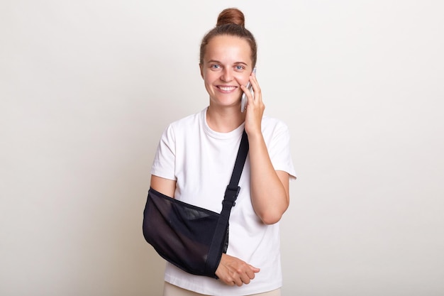 Retrato de una mujer sonriente con un peinado de moño que usa una camiseta casual de pie en un cabestrillo aislado en un fondo gris claro hablando a través de un teléfono inteligente expresando felicidad