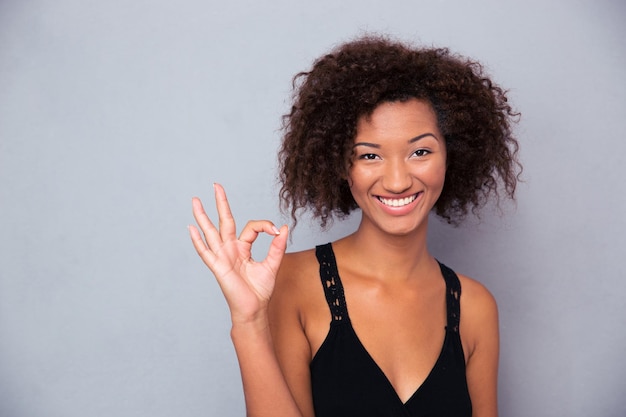 Retrato de una mujer sonriente que muestra el signo de ok con los dedos sobre la pared gris y mirando al frente