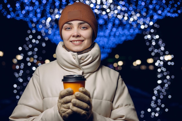Retrato de mujer sonriente en ropa exterior con taza de café calentándose las manos en la ciudad de invierno con guirnaldas