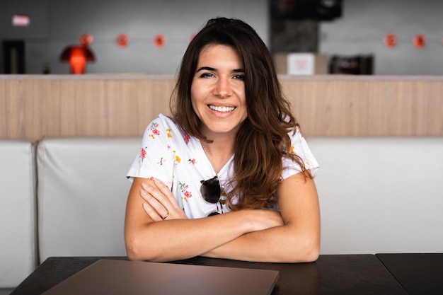 Retrato mujer sonriente con ropa informal sentada en la mesa con los brazos cruzados mirando a la cámara