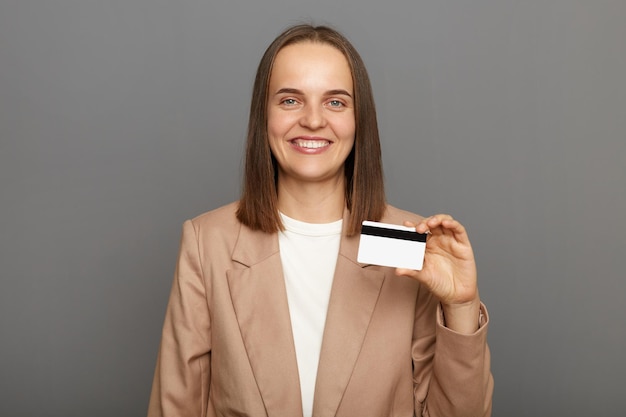 Retrato de una mujer sonriente satisfecha y optimista con el pelo castaño y una chaqueta beige posando aislada sobre un fondo gris mostrando una tarjeta de crédito mirando la cámara con felicidad