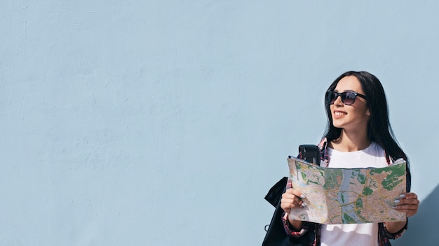 Foto retrato de mujer sonriente sosteniendo mapa de pie contra la pared azul mirando a otro lado