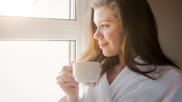 Retrato de mujer sonriente tomando café y mirando por la ventana por la mañana