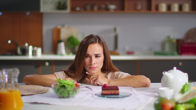 Retrato de mujer sonriente tomando decisiones entre ensalada y pastel en la cocina