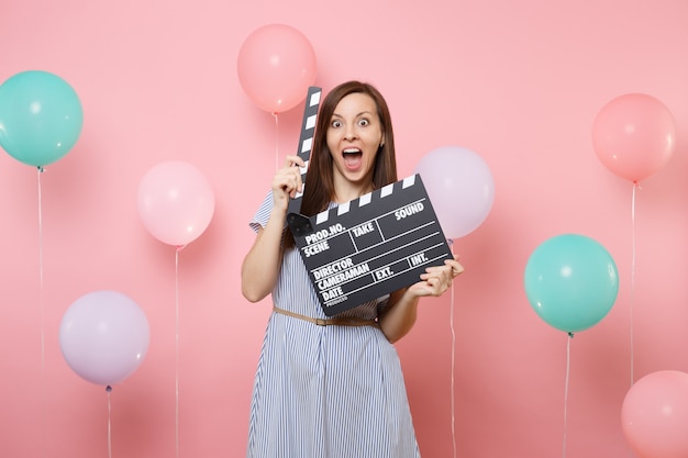 Retrato de mujer sorprendida con la boca abierta en vestido azul sosteniendo claqueta de cine negro clásico sobre fondo rosa con coloridos globos de aire. Fiesta de cumpleaños personas emociones sinceras.