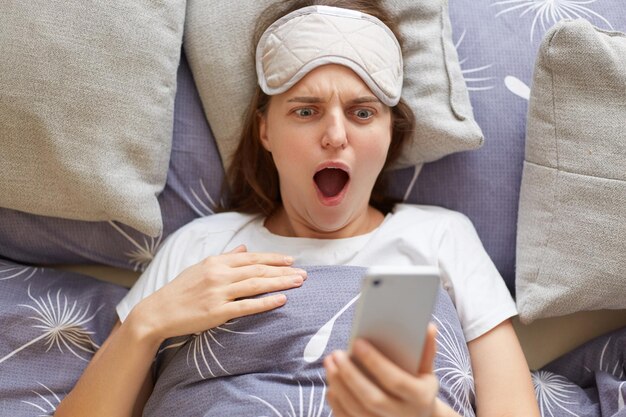 Retrato de mujer sorprendida en la cama con una máscara para dormir se despierta y mira la pantalla del teléfono usando un teléfono inteligente temprano en la mañana o en la noche gritando con asombro