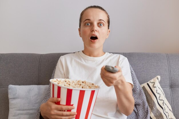 Retrato de una mujer sorprendida con una camiseta blanca de estilo casual sentada en el sofá y sosteniendo un cubo de papel con palomitas de maíz viendo una película de terror sola mantiene la boca abierta