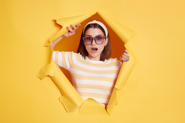 Retrato de una mujer sorprendida con una camiseta a rayas mirando a la cámara con la boca abierta que expresa asombro ve algo impactado de pie a través de un agujero roto en papel amarillo