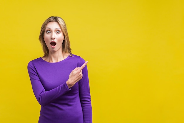 Retrato de una mujer sorprendida con un elegante vestido morado de pie apuntando a un espacio de copia vacío para información de contenido publicitario en un estudio interior aislado en un fondo amarillo