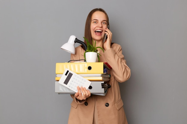 Retrato de una mujer sorprendida y emocionada con una chaqueta beige sosteniendo carpetas de papel posando aisladas sobre un fondo gris hablando por teléfono celular contando sobre su nuevo trabajo sosteniendo sus cosas