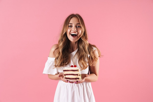 Retrato de mujer sorprendida feliz con un vestido blanco sonriendo a la cámara y sosteniendo un trozo de pastel aislado sobre la pared rosa