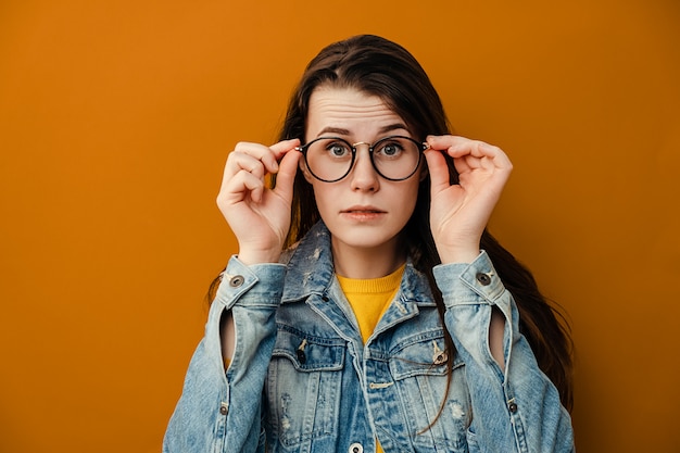 El retrato de la mujer sorprendida mantiene las manos en el borde de las gafas, se ve con expresión de omg jadea por el shock, escucha noticias asombrosas increíbles, usa una chaqueta vaquera, aislada sobre fondo marrón