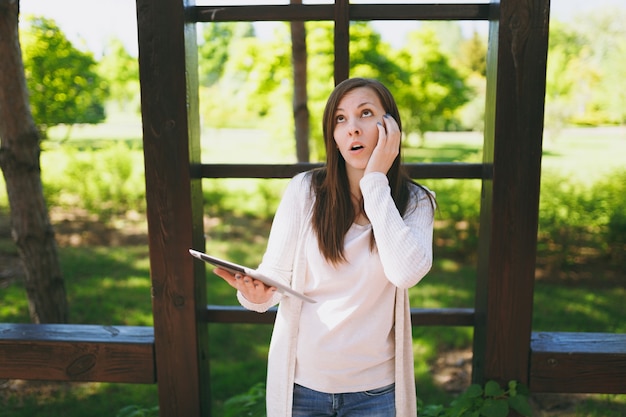 Retrato de mujer sorprendida molesta triste con ropa casual ligera. Chica guapa con tablet pc, leyendo noticias falsas en el parque de la ciudad en la calle al aire libre en primavera verde naturaleza. Concepto de estilo de vida.