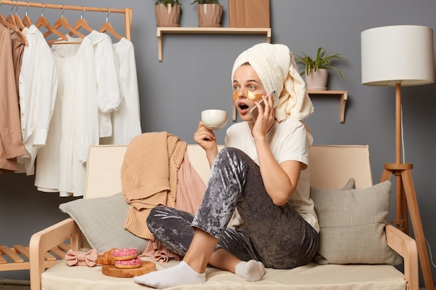 Retrato de una mujer sorprendida con pantalones grises de camiseta blanca y con una toalla en la cabeza sentada tosiendo en casa hablando por teléfono celular escuchando chismes mantiene la boca abierta