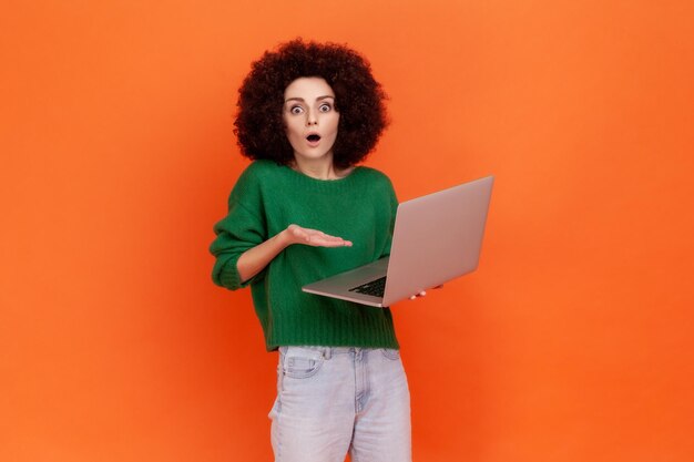 Retrato de una mujer sorprendida con peinado afro usando suéter verde de estilo casual sosteniendo un cuaderno, ve información impactante, mantiene la boca abierta. Disparo de estudio interior aislado sobre fondo naranja.