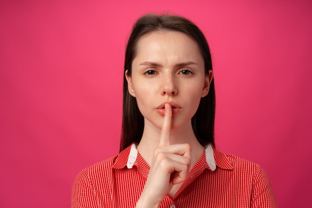 Retrato de mujer sosteniendo el dedo en los labios para guardar silencio contra el fondo de color rosa