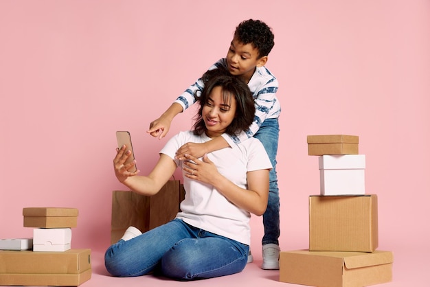 Retrato de mujer con su hijo haciendo pedidos en línea por teléfono sentados alrededor de cajas aisladas sobre rosa