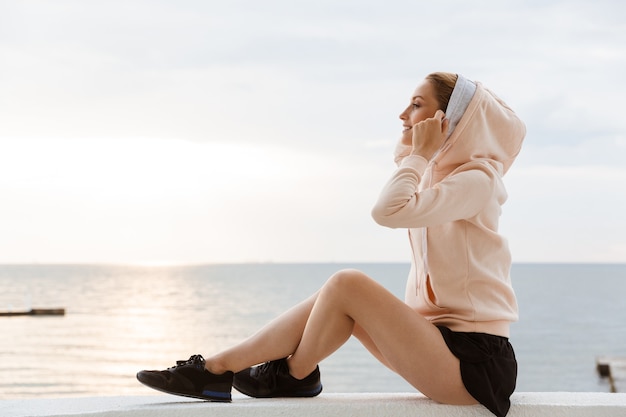 Retrato de mujer con sudadera con capucha sonriendo y mirando a un lado mientras está sentado en el muelle cerca de la playa en la mañana