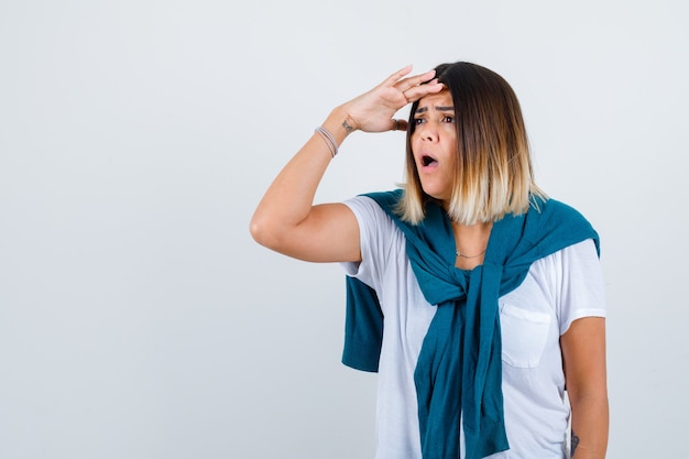 Retrato de mujer con suéter atado mirando lejos con las manos sobre la cabeza en camiseta blanca y mirando sorprendido vista frontal