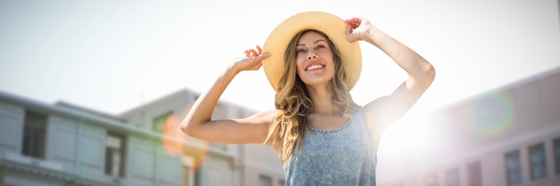 Retrato de una mujer tocando su sombrero de paja contra la vista de ángulo bajo de los edificios de la ciudad en un día soleado