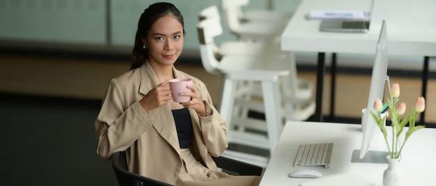 Retrato de mujer trabajadora de oficina sonriendo y mirando a la cámara mientras se relaja con una taza de bebida en el lugar de trabajo