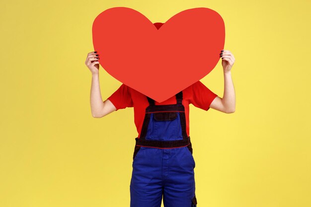 Retrato de una mujer trabajadora optimista que oculta su rostro detrás de un gran corazón rojo que expresa sentimientos románticos con un mono y una gorra roja Foto de estudio interior aislada en fondo amarillo