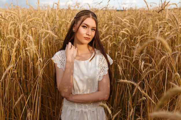 Retrato de una mujer tranquila con el pelo largo y rubio vestido de blanco de pie en un campo de trigo en verano