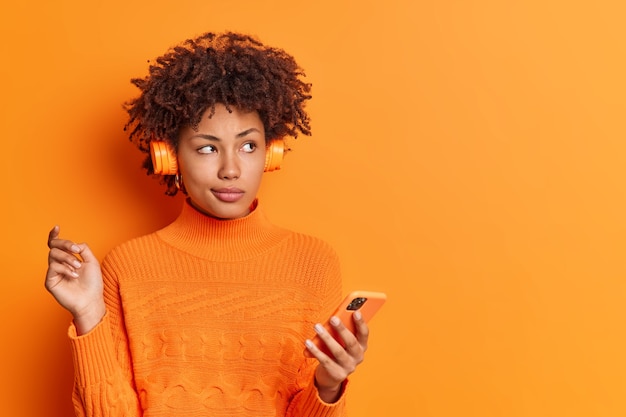 Retrato de mujer tranquila pensativa con cabello rizado usa teléfono móvil y auriculares estéreo disfruta de la lista de reproducción favorita usa un puente casual aislado sobre una pared naranja