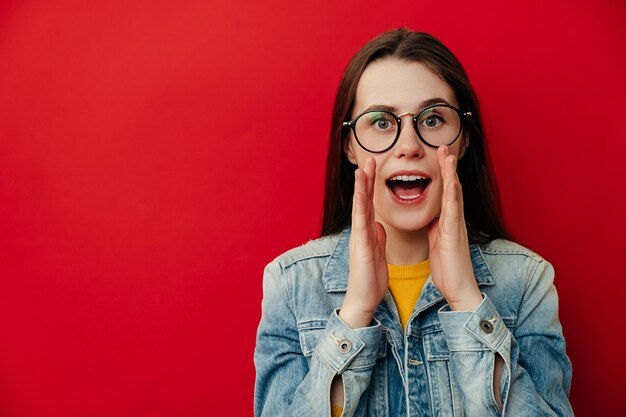 El retrato de la mujer triguena joven grita de la emoción, mantiene las manos cerca de la boca, usa la chaqueta y las gafas de mezclilla, posando aislada sobre fondo rojo. Concepto de estilo de vida de las personas. Mock up copia espacio.