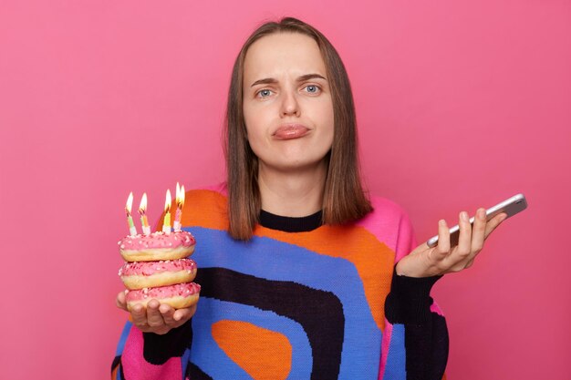 Retrato de una mujer triste con el pelo castaño usando un puente elegante sosteniendo un donut con velas y un teléfono móvil esperando a sus invitados mirando con labios de puchero aislados sobre un fondo rosa