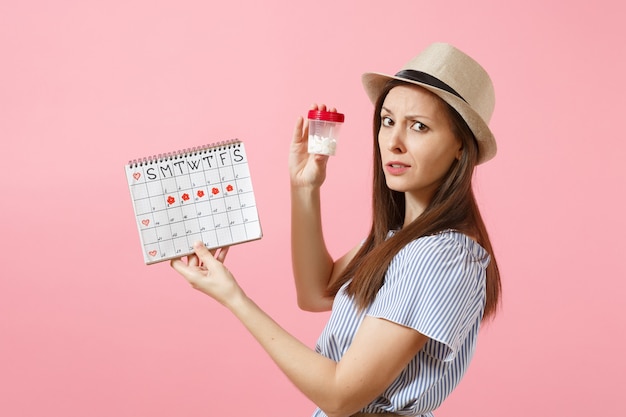 Retrato de mujer triste en vestido azul sosteniendo una botella blanca con píldoras, calendario de períodos femeninos, comprobando los días de menstruación aislados en el fondo. Atención médica, concepto ginecológico. Copie el espacio.