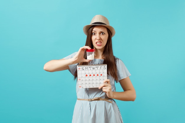 Retrato de mujer triste en vestido sosteniendo una botella blanca con píldoras, calendario de períodos femeninos, comprobando los días de menstruación aislados sobre fondo azul. Atención médica, concepto ginecológico. Copie el espacio.