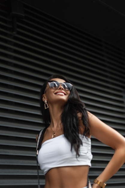 Retrato de mujer urbana feliz de una hermosa chica de moda con una sonrisa con gafas de sol en un top blanco de moda con una bolsa camina en la calle cerca de una pared de metal negro