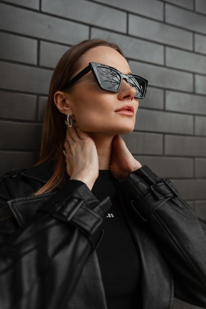 Retrato de mujer urbana de moda de una mujer hermosa y elegante con gafas de sol de moda que usa ropa negra de moda con una camiseta y una chaqueta de cuero se encuentra cerca de una pared de ladrillo negro en la calle