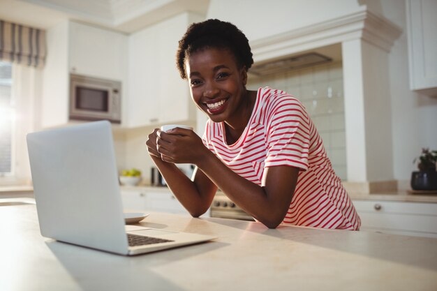 Retrato de mujer usando laptop mientras tomando un café en la cocina