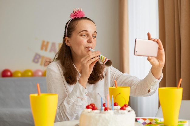 Retrato de una mujer vestida de blanco sentada en la mesa con pastel y bebida, sosteniendo un teléfono móvil y haciendo una videollamada, haciendo sonar la fiesta en línea.