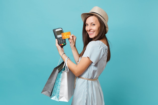 Retrato de mujer vestida, sombrero sosteniendo bolsas de paquetes con compras después de ir de compras, terminal de pago bancario moderno inalámbrico para procesar y adquirir pagos con tarjeta de crédito aislados sobre fondo azul pastel.