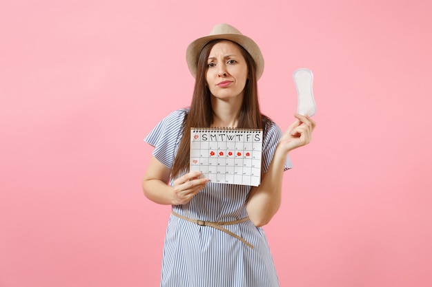 Retrato de mujer con vestido azul, sombrero con toalla sanitaria, calendario de períodos femeninos para comprobar los días de menstruación aislados sobre fondo rosa. Concepto médico, sanitario, ginecológico. Copia espacio