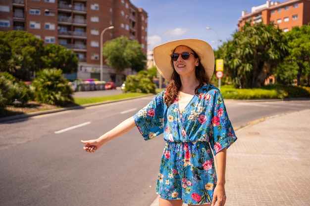 Retrato de una mujer con vestido floral de verano y sombrero haciendo autostop junto a una calle