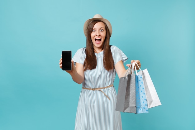 Retrato de mujer en vestido de verano, sombrero de paja con bolsas de paquetes con compras después de ir de compras, teléfono móvil con pantalla vacía aislada sobre fondo azul pastel. Copie el espacio para publicidad.