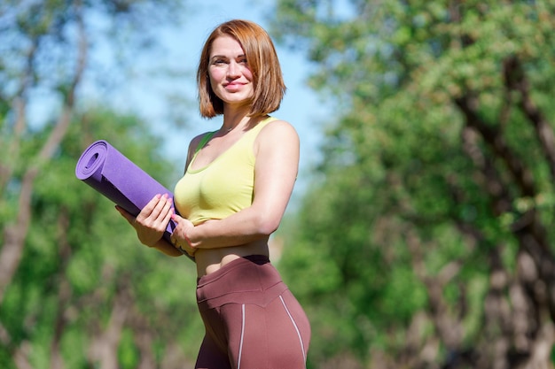 Retrato de mujer de yoga con alfombra púrpura en manos en el bosque en verano